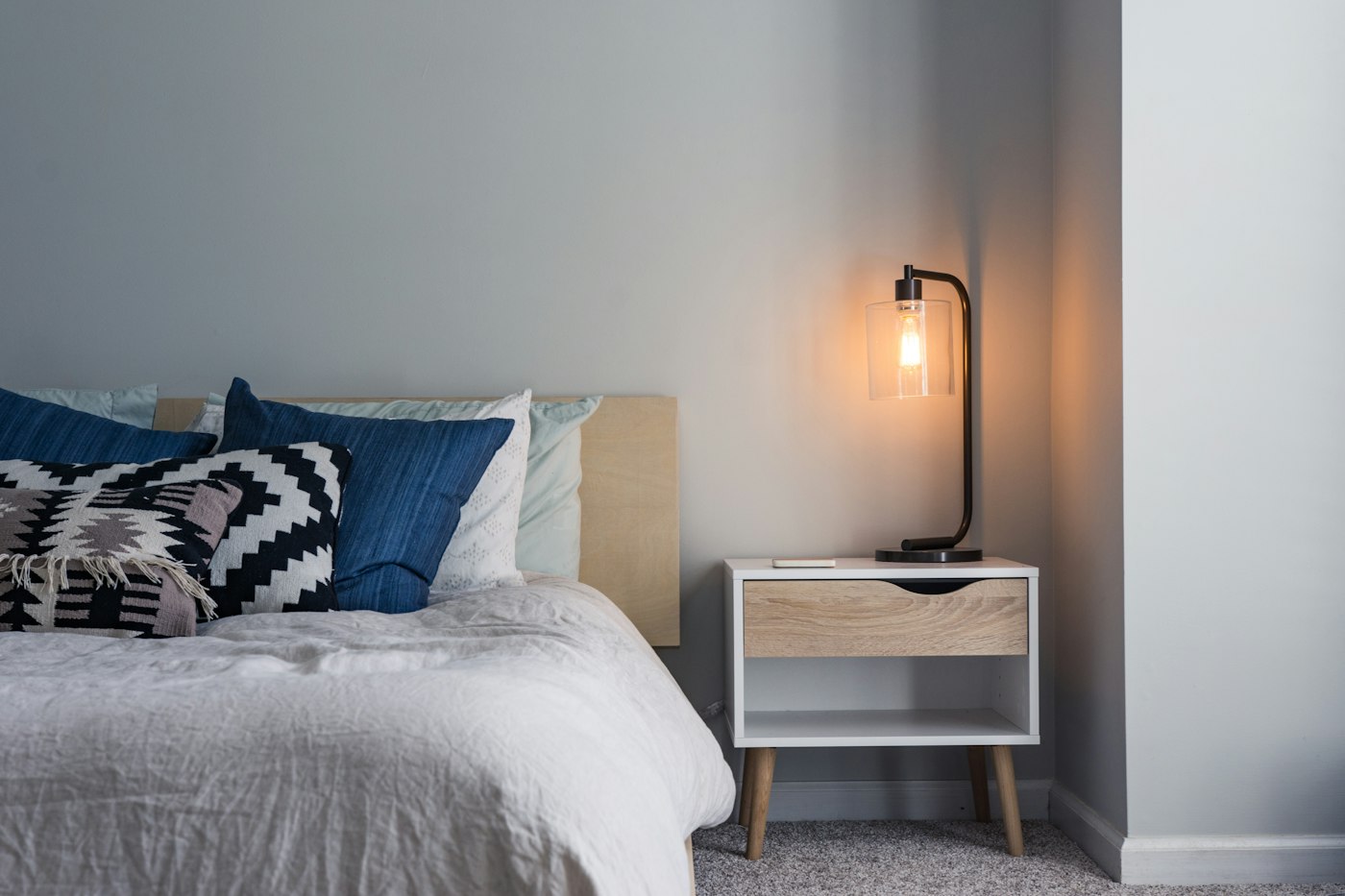 Bed with a white cover, blue cushions, a patterned cushion, and a lit lamp on a small cabinet.